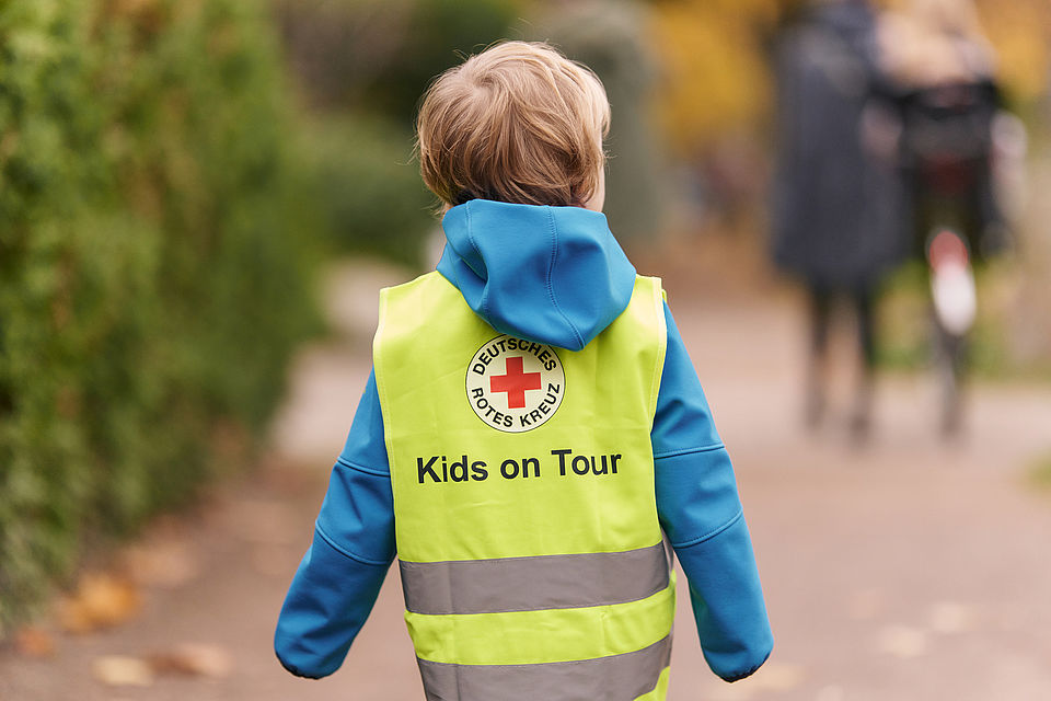 Ein Bild eines Kindes vin hinten. Das Kind hat eine gelbe Warnweste mit dem DRK Logo an. Kids on tour steht darauf.