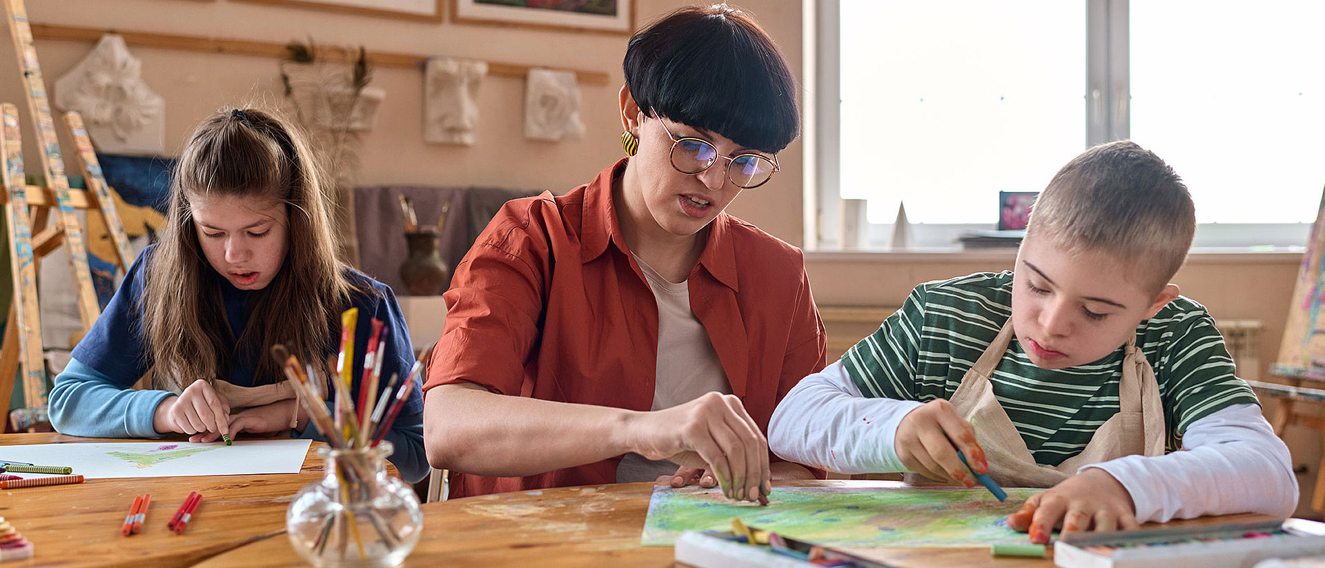 Drei Personen malen an einem Tisch. Auf dem Tisch liegen Pinsel und Farben. Im Hintergrund hängen Bilder und stehen Staffeleien.