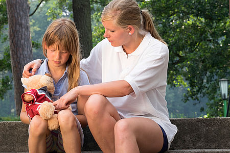 Eine Frau die Ein Kind tröstet. Das Kind hält einen DRK-Teddy in der Hand und guckt traurig.