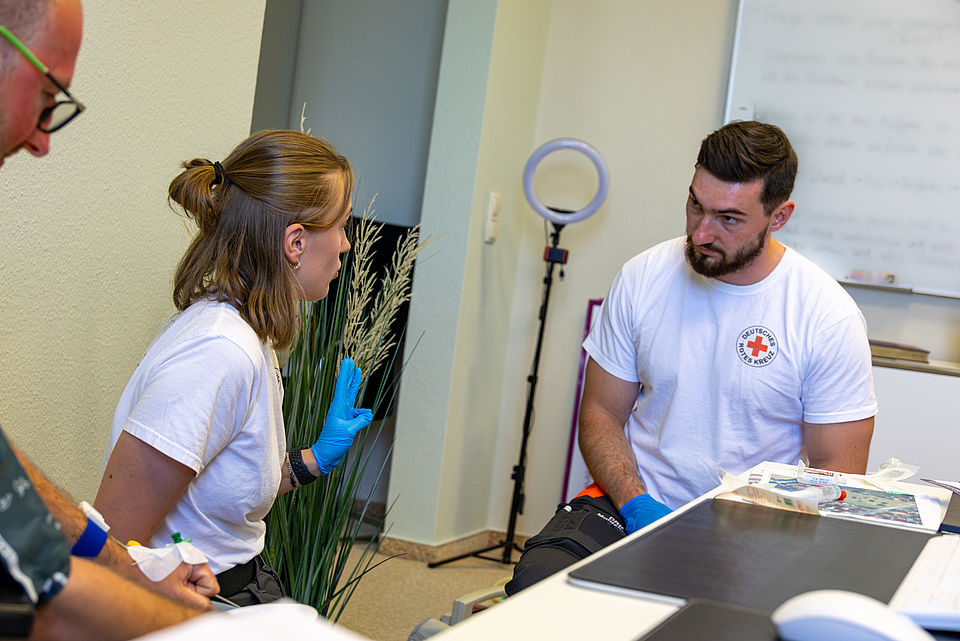 Eine junge Frau in einem weißen T-Shirt des Deutschen Roten Kreuzes (DRK) und blauen Handschuhen spricht gestikulierend mit einem jungen Mann, der ebenfalls ein DRK-T-Shirt trägt. Beide hocken während des Gespräches.