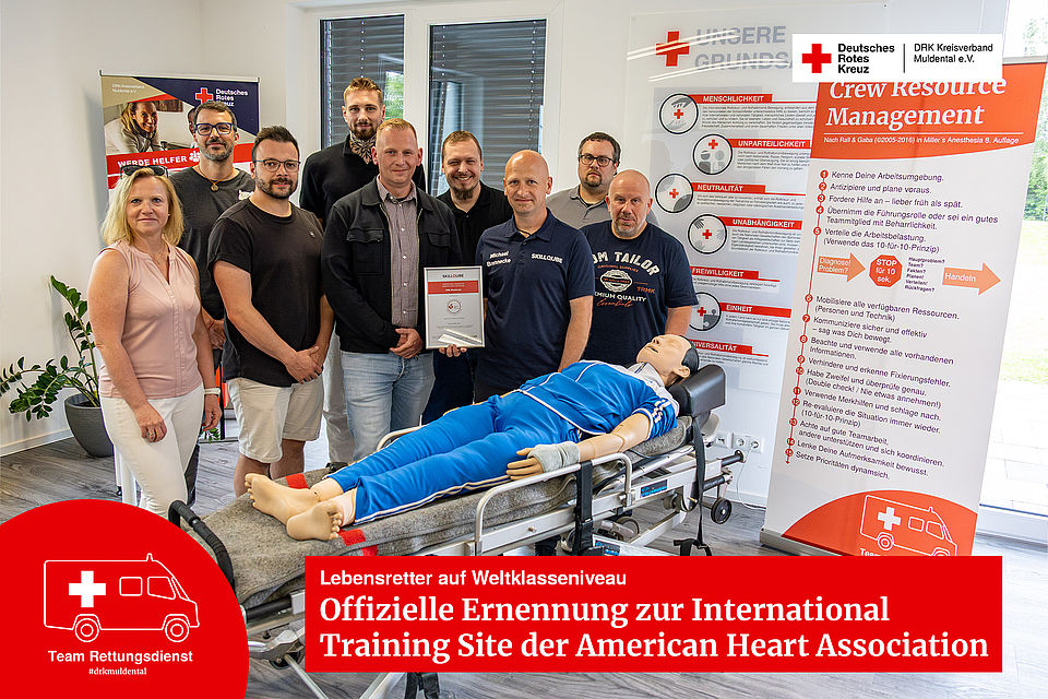 Gruppenbild des DRK-Rettungsdienst-Teams zur AHA-Zertifizierung. Zehn Beteiligte stehen in einem hellen Schulungsraum hinter einer medizinischen Simulationspuppe auf einer Krankentrage. In der Mitte wird das frisch überreichte Zertifikat präsentiert.