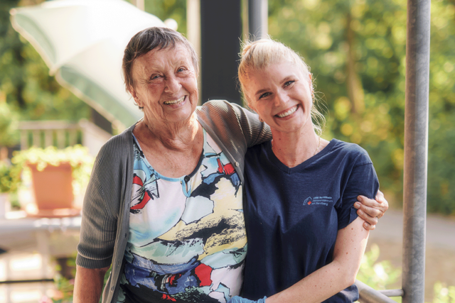 Eine jugen Frau stützt eine ältere Dame beim Gehen. Beide lächeln in die Kamera.