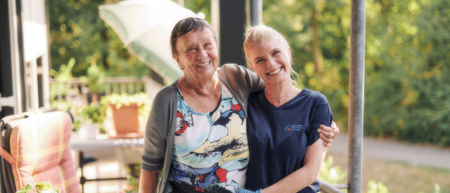 Eine jugen Frau stützt eine ältere Dame beim Gehen. Beide lächeln in die Kamera.