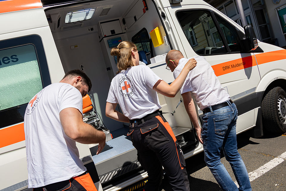 Eine junge Frau in einem weißen DRK-T-Shirt und Einsatzhose hilft einem Mann in einem weißen Hemd und Jeans, aus einem weißen Krankenwagen auszusteigen. Ein weiterer Mann in einem DRK-T-Shirt und orangefarbener Hose steht links vor dem Krankenwagen und scheint etwas vorzubereiten. Das Heck des Krankenwagens ist geöffnet, und die Aufschrift "DRK Muldental" ist sichtbar.