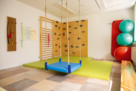 Ein heller Therapieraum mit einer hängenden blauen Therapieschaukel über grünen Matten, einer Sprossen- und Kletterwand sowie Gymnastikbällen.