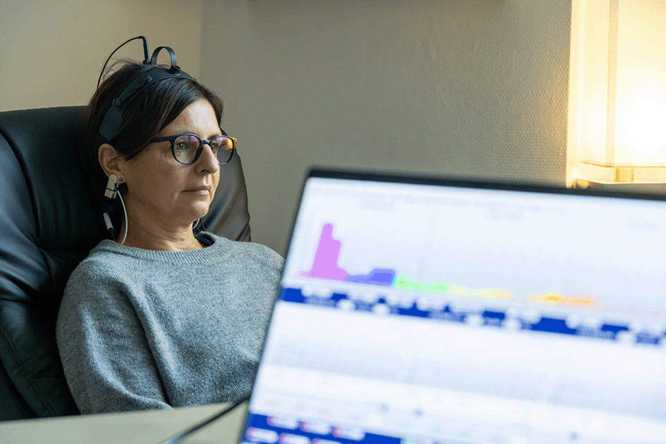 Neurofeedback Symbolbild Eine Frau in einem Sessel trägt Sensoren am Kopf und Ohrclips für eine Neurofeedback-Therapie, während sie konzentriert auf einen Computerbildschirm blickt. Vom Vordergrund ist unscharf ein Bildschirm mit grafischen Auswertungen ihrer Gehirnwellen.