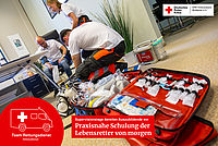 Zwei junge Sanitäter in Ausbildung knien vor einer geöffneten roten Notfalltasche mit medizinischen Geräten, während im Hintergrund ein Mann von einem anderen Sanitäter den Blutdruck gemessen bekommt.