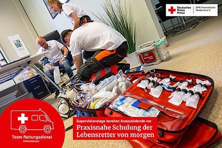 Zwei junge Sanitäter in Ausbildung knien vor einer geöffneten roten Notfalltasche mit medizinischen Geräten, während im Hintergrund ein Mann von einem anderen Sanitäter den Blutdruck gemessen bekommt.