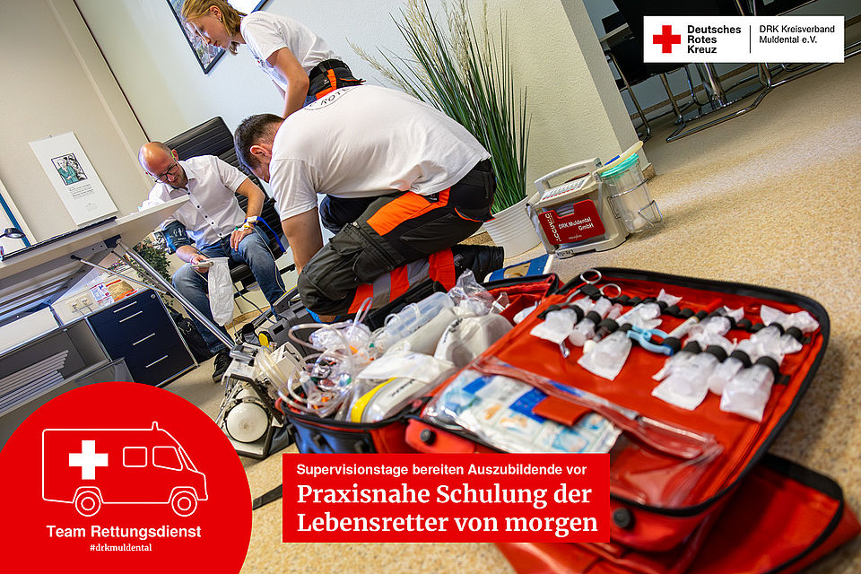 Zwei junge Sanitäter in Ausbildung knien vor einer geöffneten roten Notfalltasche mit medizinischen Geräten, während im Hintergrund ein Mann von einem anderen Sanitäter den Blutdruck gemessen bekommt.