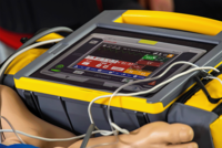 Eine Nahaufnahme eines medizinischen Defibrillators vom Typ ‚Schiller Defigard Touch 7‘ während eines Einsatzes oder Trainings. Das Display zeigt detaillierte Vitalparameter und Einstellungen wie den Energielevel von 200 Joule an.