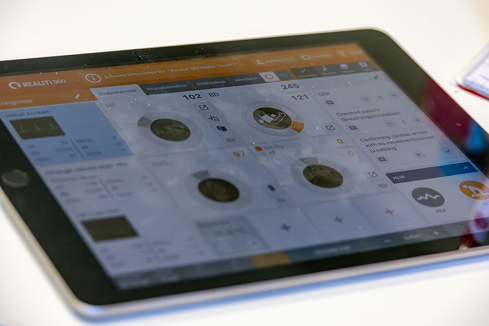 Nahaufnahme eines Tablets, das die Benutzeroberfläche einer medizinischen Simulationssoftware namens "REALITI360" anzeigt. Auf dem Bildschirm sind Vitalparameter wie Blutdruck (102/80), Herzfrequenz (121) und weitere medizinische Daten sowie eine Checkliste oder Notizen zu sehen.