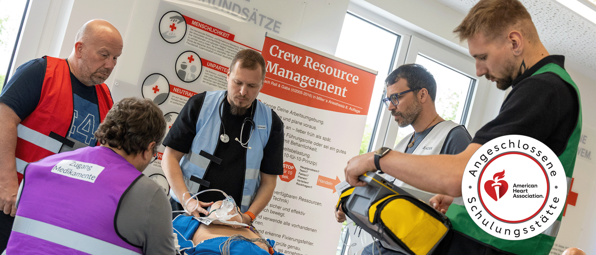 ACLS Landingpage header Ein Team aus vier Einsatzkräften führt ein strukturiertes Training in einer medizinischen Schulung durch. Ein Teilnehmer in einer violetten Weste mit der Aufschrift ‚Zugang Medikamente‘ assistiert bei der Versorgung einer Übungspuppe, während ein Teammitglied in einer hellblauen Weste mit Stethoskop konzentriert die Beatmung durchführt. Im Hintergrund stehen Roll-ups zu den DRK-Grundsätzen und zum Thema ‚Crew Resource Management‘. Rechts im Bild hält ein Teilnehmer einen Defibrillator bereit. Das Bild trägt das offizielle Siegel der ‚Angeschlossenen Schulungsstätte der American Heart Association