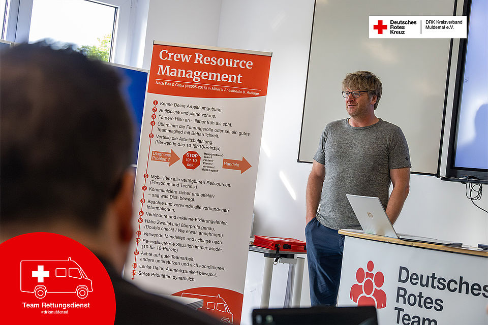 Blick über die Schulter eines Teilnehmers auf den ärztlichen Leiter Rettungsdienst Robert Stöhr, der im DRK-Schulungsraum einen Vortrag hält. Neben ihm ist ein Aufsteller zum Thema „Crew Resource Management“ platziert.
