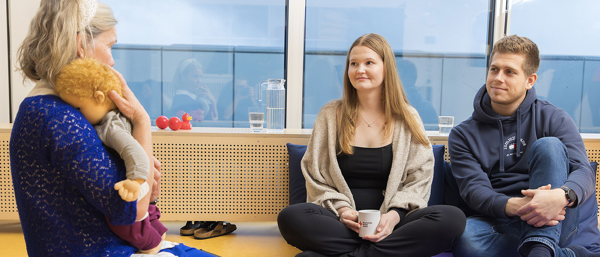 Baby - Beratung im DRK-Klinikum Westend in Berlin Eine Gruppe von drei Personen sitzt in einem hellen Raum. Eine Frau auf der linken Seite hält eine Babypuppe im Arm und spricht mit einer jungen Frau und einem jungen Mann. Beide sitzen auf dem Boden und schauen aufmerksam zur Frau mit der Puppe.