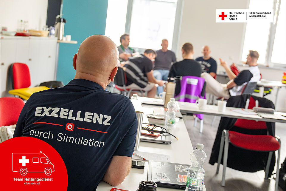 Nahaufnahme eines Poloshirts mit dem Aufdruck „EXZELLENZ durch Simulation“. Im unscharfen Hintergrund ist eine Schulungsgruppe des DRK-Rettungsdienstes zu sehen, die in einer Diskussionsrunde zusammensitzt.