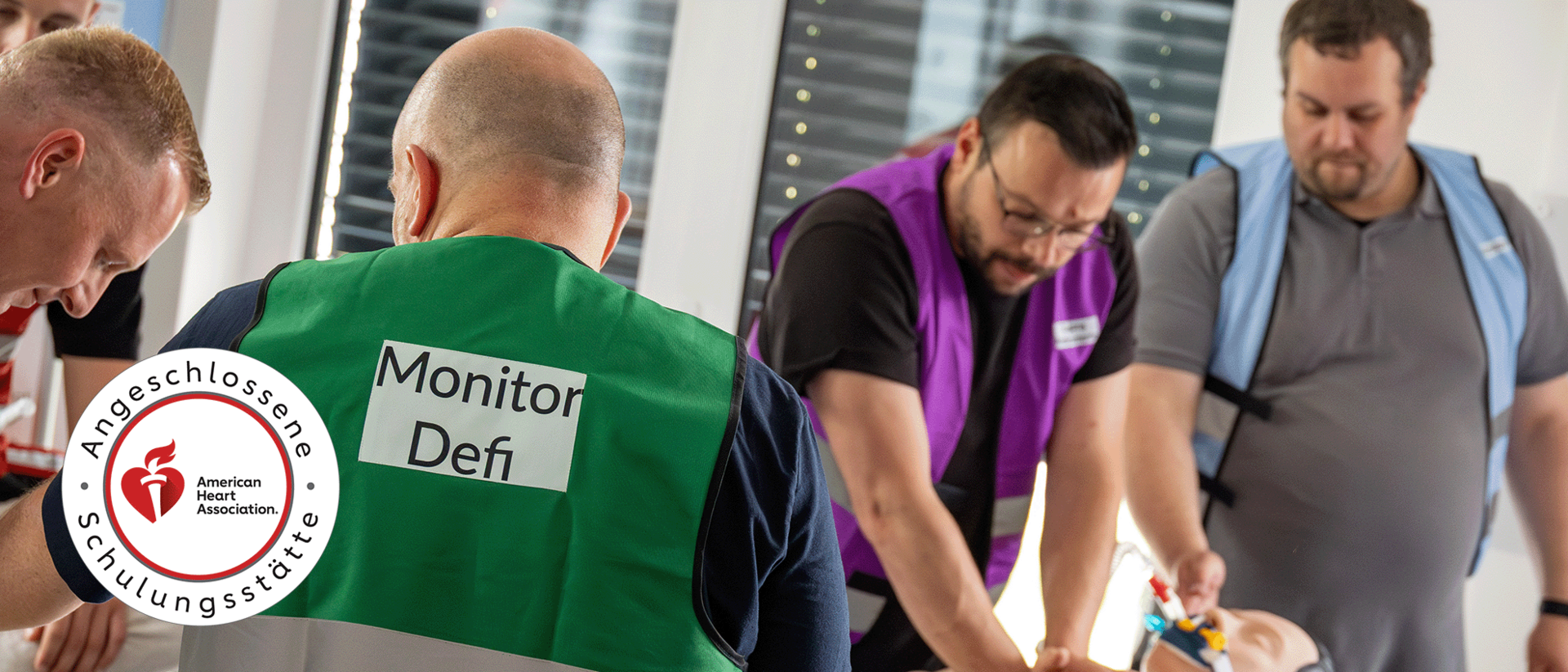 ACLS Refresher header In einem medizinischen Schulungsraum beugen sich vier Teilnehmer über eine Übungspuppe, um Herz-Lungen-Wiederbelebung zu trainieren. Zwei Männer im Vordergrund tragen grüne und violette reflektierende Warnwesten, die ihre spezifischen Aufgaben als ‚Monitor/Defi‘ und Teamleiter kennzeichnen. Ein dritter Teilnehmer in einer hellblauen Weste assistiert, während der vierte von links den Kopf der Puppe hält. Links unten ist das Siegel der ‚Angeschlossenen Schulungsstätte der American Heart Association‘ zu sehen
