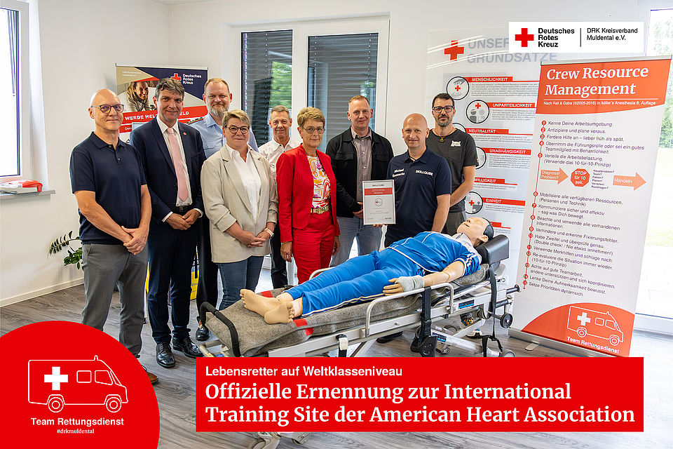 Gruppenfoto zur offiziellen AHA-Zertifizierung beim DRK Muldental. Neun Projektbeteiligte und Führungskräfte posieren lächelnd um eine medizinische Reanimations-Übungspuppe, die auf einer Rettungsdienst-Trage liegt. Ein Beteiligter hält die gerahmte Zertifikatsurkunde in die Kamera.