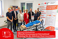 Gruppenfoto zur offiziellen AHA-Zertifizierung beim DRK Muldental. Neun Projektbeteiligte und Führungskräfte posieren lächelnd um eine medizinische Reanimations-Übungspuppe, die auf einer Rettungsdienst-Trage liegt. Ein Beteiligter hält die gerahmte Zertifikatsurkunde in die Kamera.