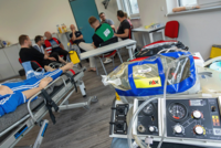 Ein Einblick in eine ACLS-Präsenzschulung: Im Vordergrund steht medizinische Ausrüstung wie ein Beatmungsgerät und eine Notfalltasche mit der Aufschrift ‚Airway‘. Im Hintergrund sind Teilnehmende und Instruktoren in einem Schulungsraum zu sehen, die an einer Übungspuppe arbeiten.