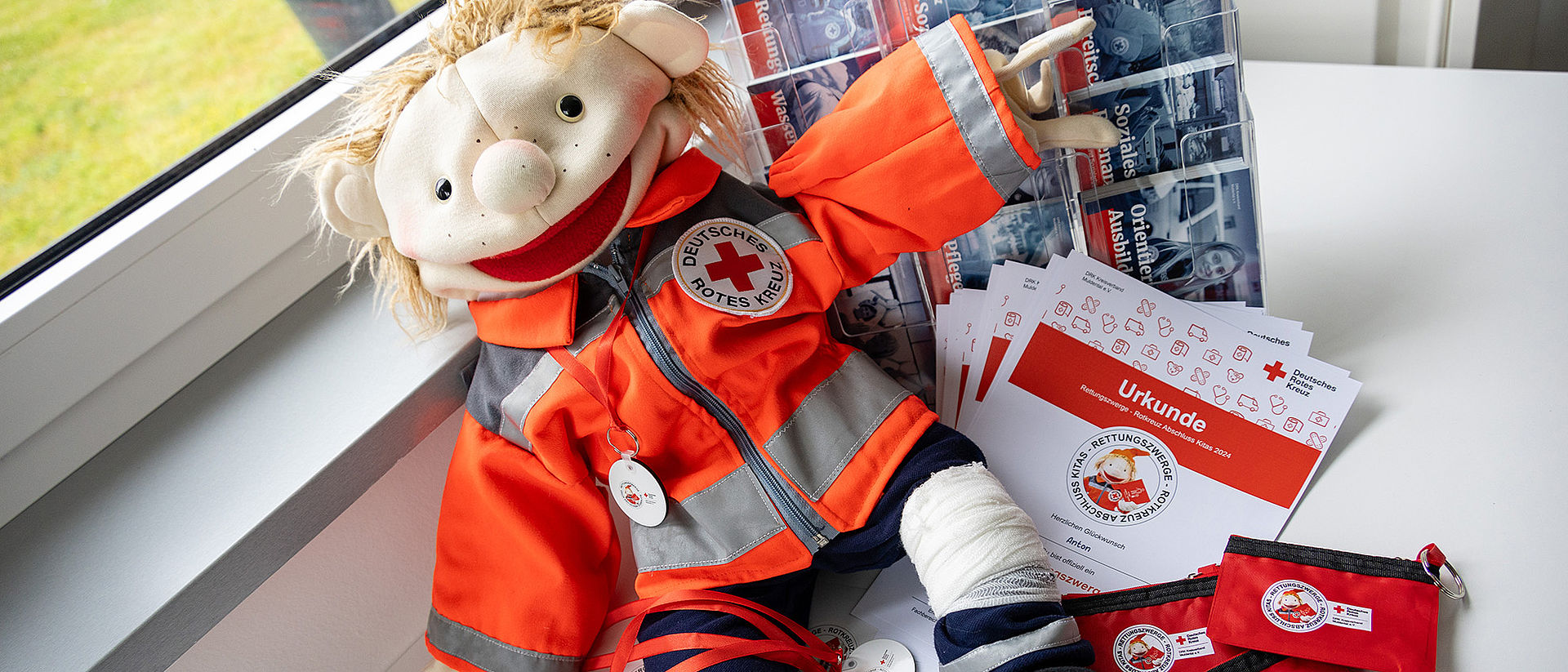 Eine Puppe in orangefarbener DRK-Uniform sitzt auf einem Tisch. Daneben liegen Urkunden, Schlüsselanhänger, rote Täschchen und Infomaterial.