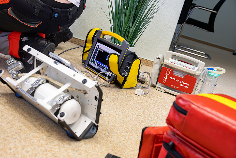 Nahaufnahme von medizinischen Geräten, die auf dem Boden liegen, darunter ein gelber Defibrillator/Monitor, ein Sauerstoff-System. Im Vordergrund sind die Beine einer Person in Sanitäterkleidung zu sehen.
