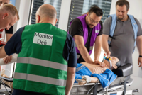 In einem medizinischen Schulungsraum beugen sich vier Teilnehmer über eine Übungspuppe, um Herz-Lungen-Wiederbelebung zu trainieren. Zwei Männer im Vordergrund tragen grüne und violette reflektierende Warnwesten, die ihre spezifischen Aufgaben als ‚Monitor/Defi‘ und Teamleiter kennzeichnen. Ein dritter Teilnehmer in einer hellblauen Weste assistiert, während der vierte von links den Kopf der Puppe hält.