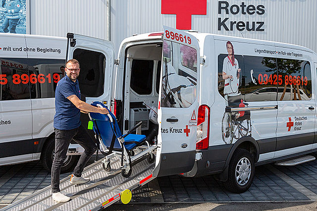 Ein Mitarbeiter des Fahrdienstes schiebt den Rollstuhl die Laderampe hoch ins Auto.