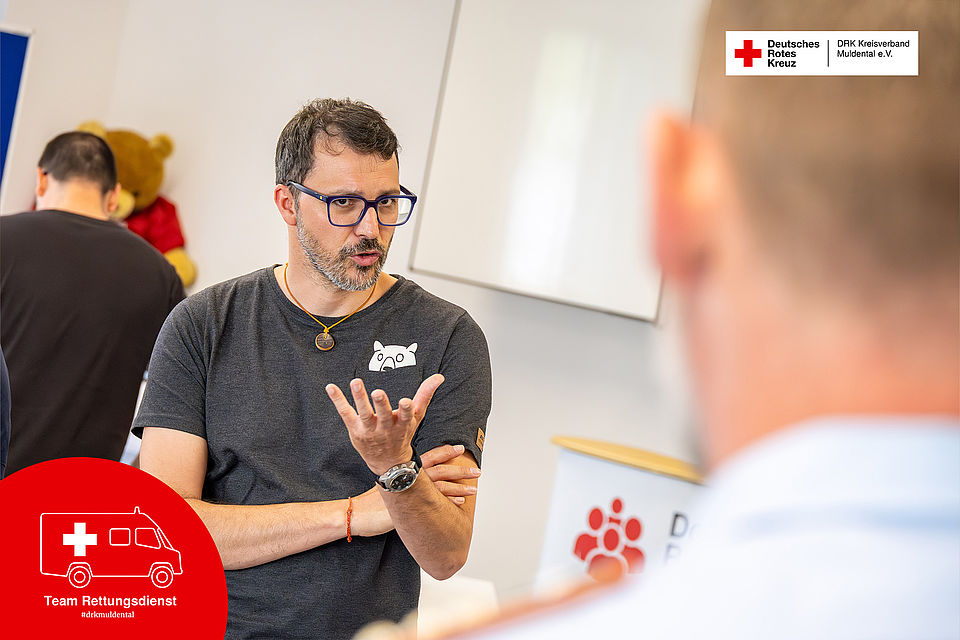 Leitender Praxisanleiter Tobias Ude des DRK-Rettungsdienstes Muldental im Gespräch während der AHA-Schulung. Er untermalt seine Erklärung mit einer Handgeste, während ein Kollege im Vordergrund unscharf im Bild steht.