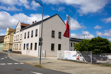 Ein modernes Gebäude mit hellerer Fassade und angrenzendem Altbau unter blauem Himmel, davor eine DRK-Flagge und ein Werbeplakat.
