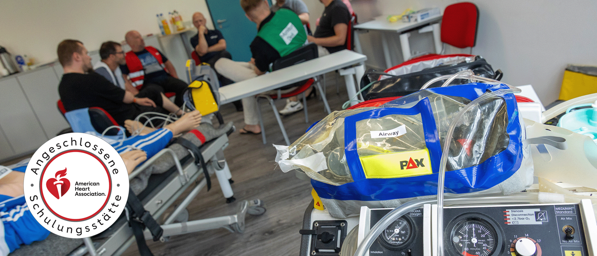 ACLS Präsenz header Ein Einblick in eine ACLS-Präsenzschulung: Im Vordergrund steht medizinische Ausrüstung wie ein Beatmungsgerät und eine Notfalltasche mit der Aufschrift ‚Airway‘. Im Hintergrund sind Teilnehmende und Instruktoren in einem Schulungsraum zu sehen, die an einer Übungspuppe arbeiten. Links ist das Logo der American Heart Association als zertifizierte Schulungsstätte zu sehen.