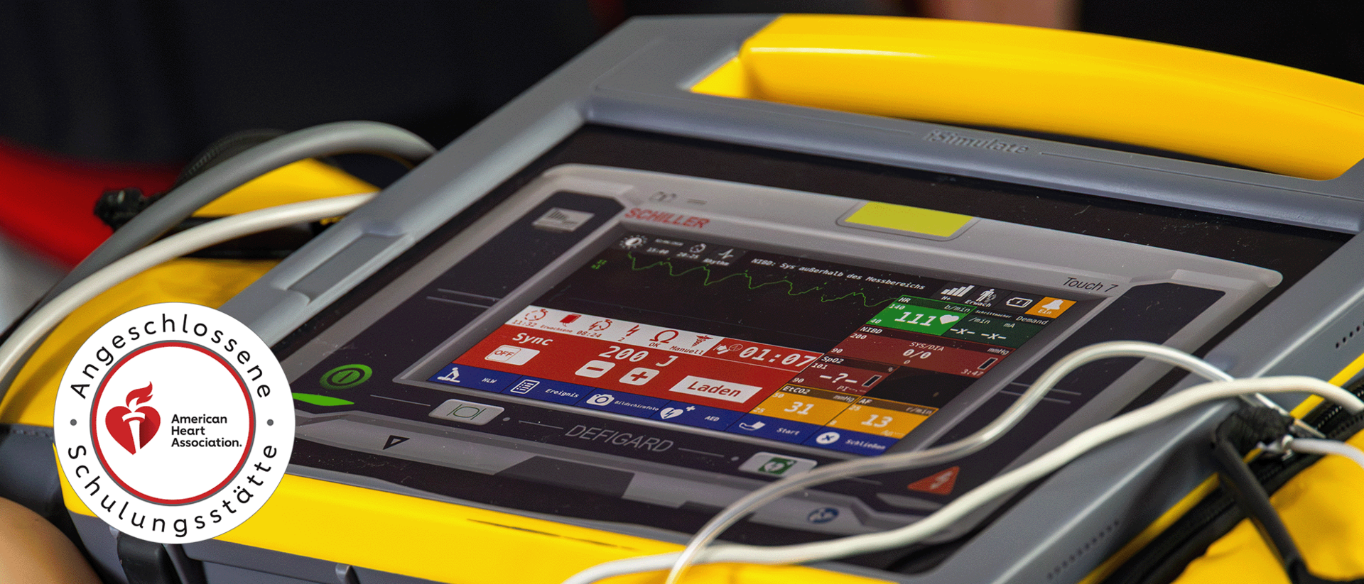 ACLS heartcode header Eine Nahaufnahme eines medizinischen Defibrillators vom Typ ‚Schiller Defigard Touch 7‘ während eines Einsatzes oder Trainings. Das Display zeigt detaillierte Vitalparameter und Einstellungen wie den Energielevel von 200 Joule an. Links im Bild ist das Siegel der ‚Angeschlossenen Schulungsstätte der American Heart Association‘ zu sehen, welches die Zertifizierung des Kurses verdeutlicht.