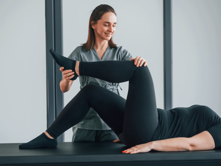 kairos Symbolbild Eine lächelnde Physiotherapeutin in grauer Arbeitskleidung behandelt eine Patientin, die auf einer Behandlungsliege liegt. Die Therapeutin führt eine Dehnübung durch, indem sie das angewinkelte Bein der Patientin, die schwarze Sportkleidung trägt, sanft mit ihren Händen hält und bewegt. Die Szene findet in einer hellen, professionellen Praxisumgebung statt.