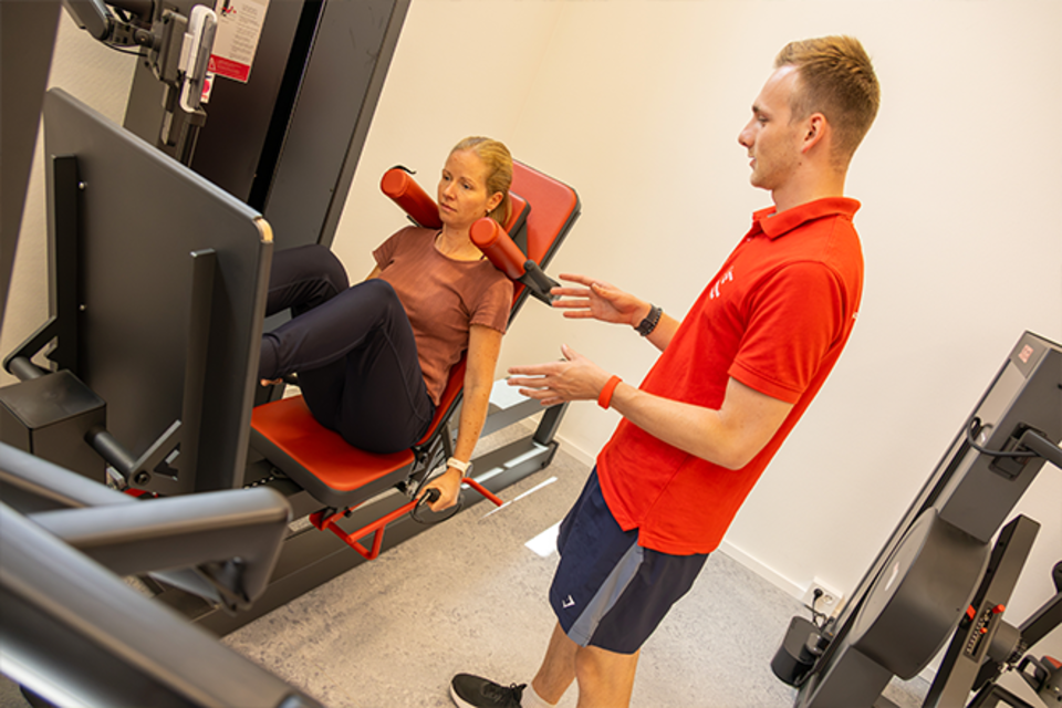 Ein Trainer im roten DRK-Shirt weist eine Frau in einem Fitnessstudio an einem medizinischen Beinpressgerät ein.